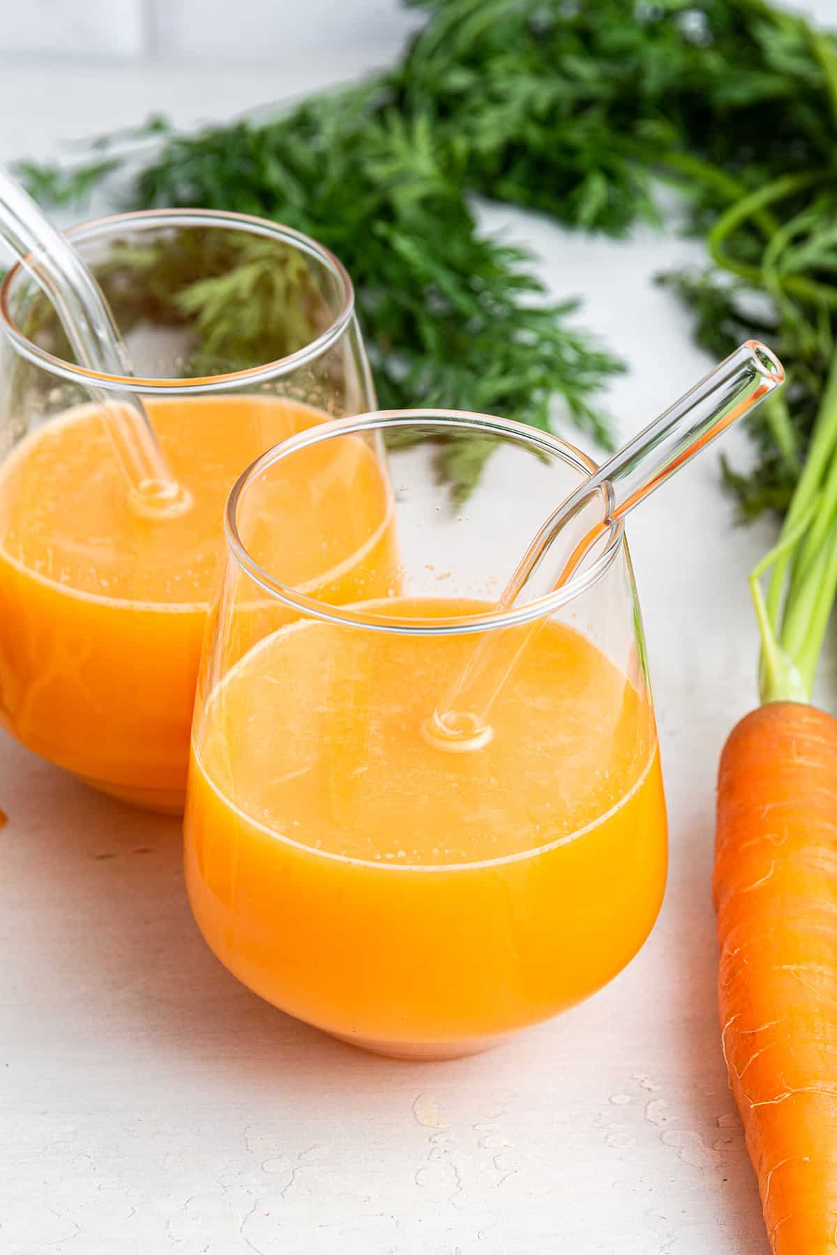 This refreshing carrot juice recipe has loads of health benefits! I’ll show you two easy ways to make it: in a juicer and in a blender. Two glasses of carrot juice with glass straws set next to carrot on counter