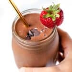 A hand holding a chocolate strawberry smoothie in a glass jar garnished with chocolate chips and a fresh berry.