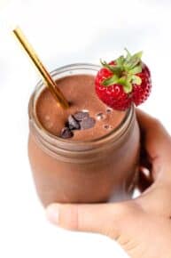 A hand holding a chocolate strawberry smoothie in a glass jar garnished with chocolate chips and a fresh berry.