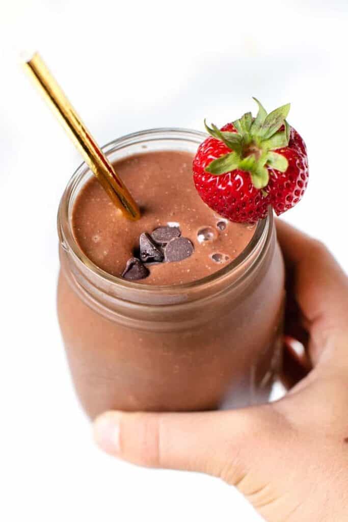 A hand holding a chocolate strawberry smoothie in a glass jar garnished with chocolate chips and a fresh berry.