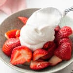 A spoon dolloping fluffy coconut whipped cream onto a bowl of strawberries.