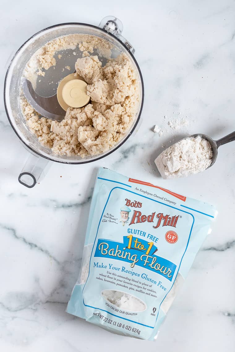 Overhead view of a gluten-free pie crust mixture inside a blender, next to a bag of 1-to-1 gluten-free baking flour.