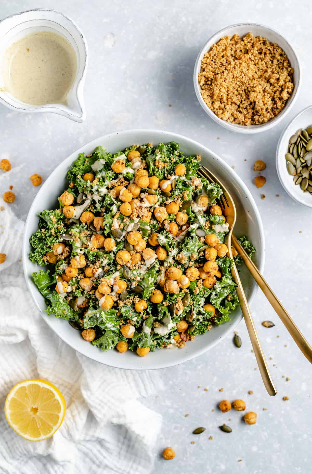 Overhead view of vegan kale Caesar salad in a white bowl topped with crispy chickpeas and tahini dressing, surrounded by salad ingredients.