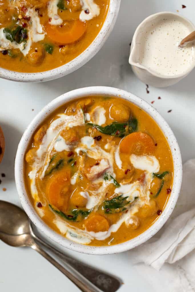 Overhead view of a bowl of chickpea and pumpkin stew swirled with vegan cream, next to a spoon, a creamer jug, and a second bowl of stew.