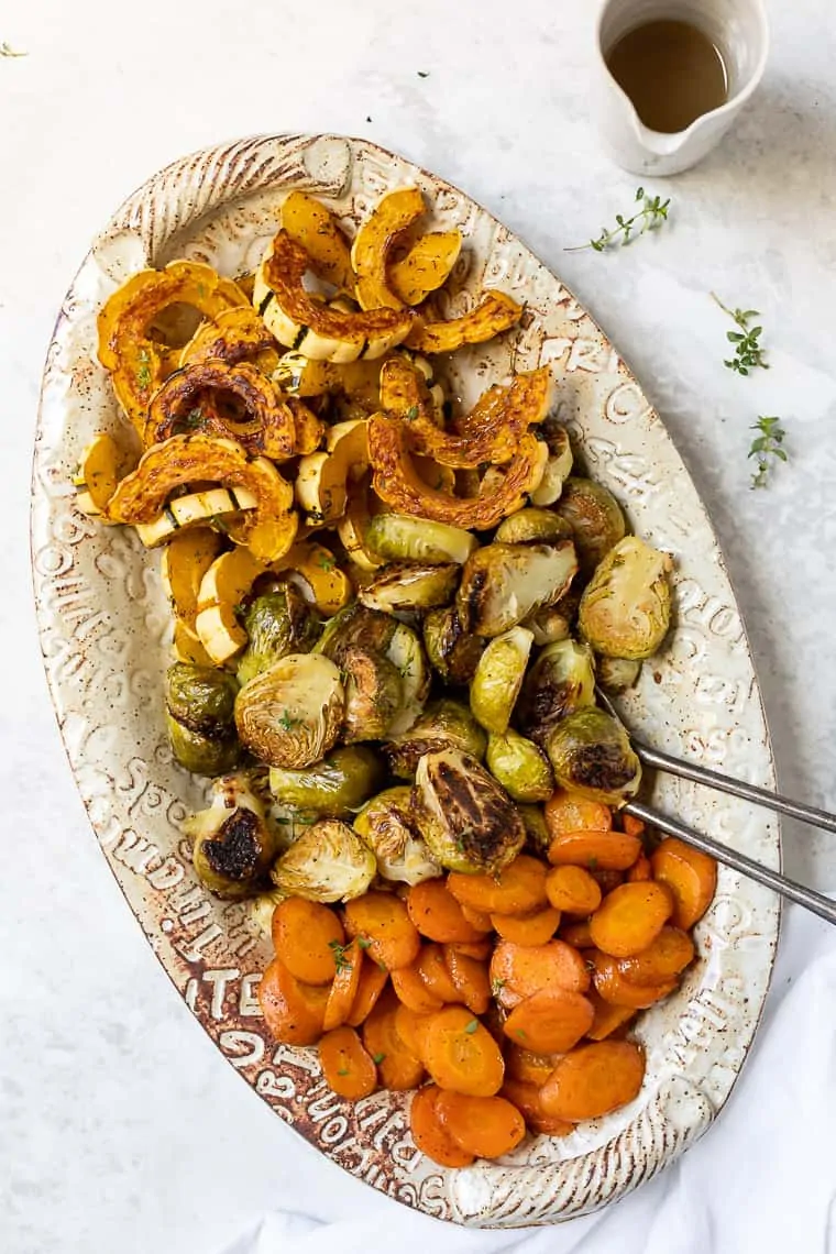 Overhead view of a platter of roasted winter vegetables, including delicata squash, Brussels sprouts, and carrots next to a small pourer of maple glaze.