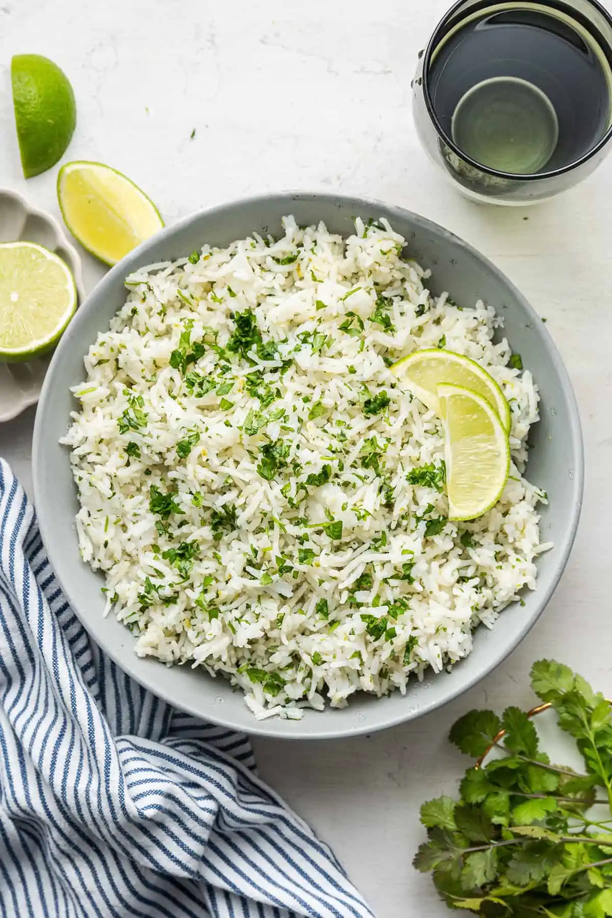This cilantro lime rice is full of fresh and fragrant lime juice and zest, and is the perfect side dish for Mexican or Asian meals. A plate of cilantro lime rice topped with lime slices, next to a sprig of cilantro, fresh limes, a kitchen towel, and a water glass