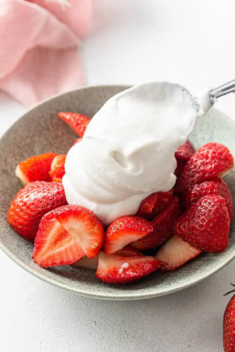 A spoon dolloping fluffy coconut whipped cream onto a bowl of strawberries.