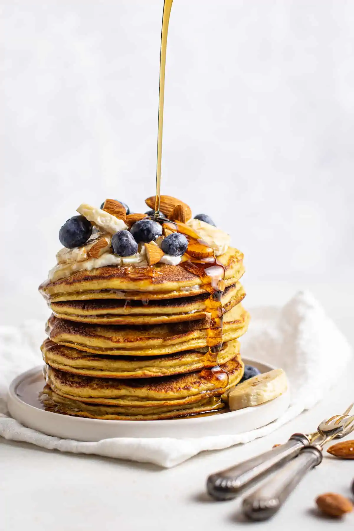 A stack of protein pancakes on a plate topped with fresh fruit and nuts, drizzled with syrup.