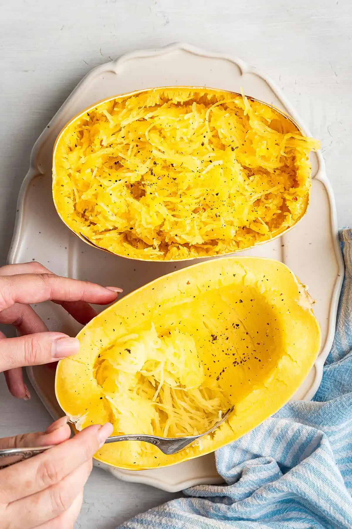 A cooked spaghetti squash cut in half on a serving tray, with one half shredded, and the other half in the process of being shredded by a fork