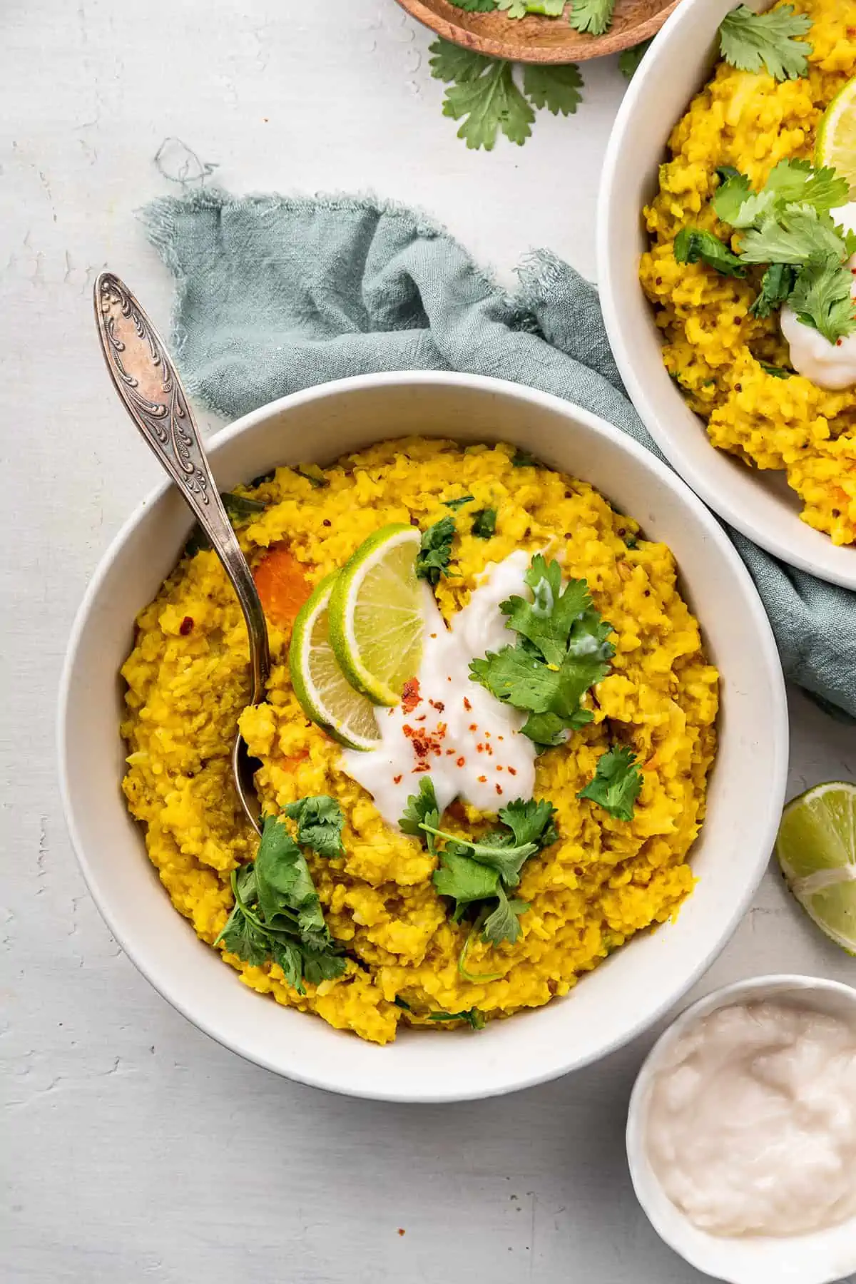 This kitchari recipe is a staple of Ayurvedic eating, and is healing for the mind, body, and soul, and excellent for your gut health. Overhead view of a bowl of kitchari topped with yogurt, cilantro, and lime slices, with a spoon in it, next to another bowl.