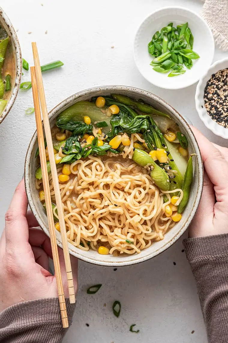 This easy vegan ramen recipe is a quick and flavorful one-pot dinner packed with tender veggies and slurpable noodles in a rich miso broth. Overhead view of a bowl of vegan ramen with hands wrapped around the bowl and chopsticks on the side.